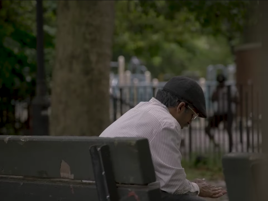 A zoomed out still image from the film 'Reflections' of a middle aged man sitting on a bench looking at the ground.